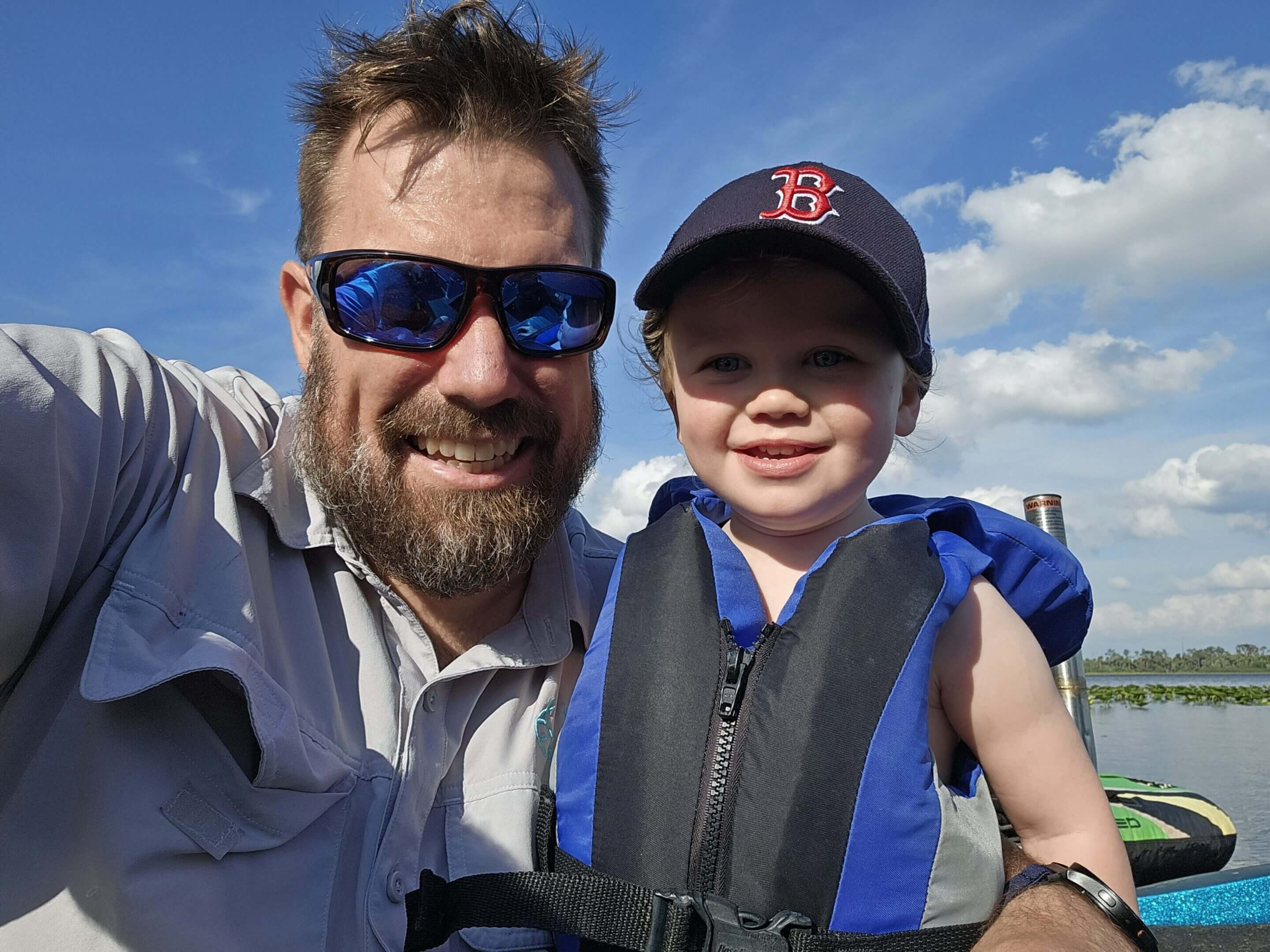 A man and a young boy smiling outdoors on a sunny day.